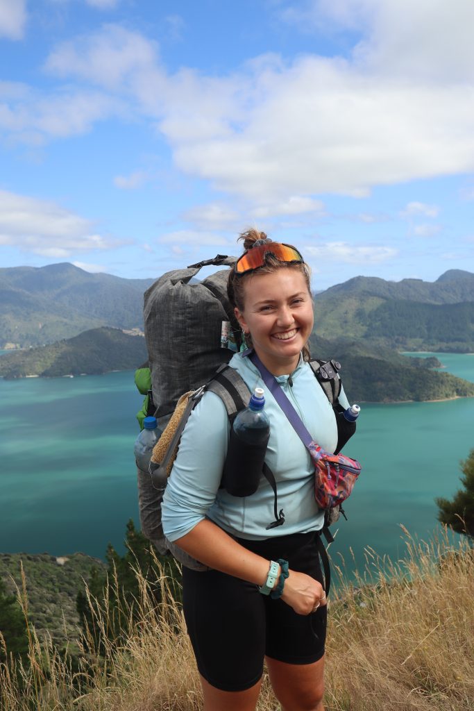 Ellie smiling with her backpack on and a beautiful New Zealand coastal landscape behind her