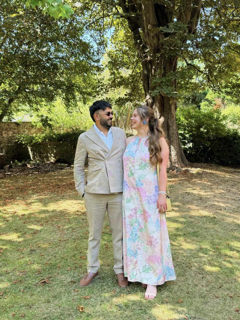 Lily with her partner Nam, they stand on grass in front of trees, dressed up, him in a beige suit and Lily in a floral floor-length dress and looking at each other smiling 