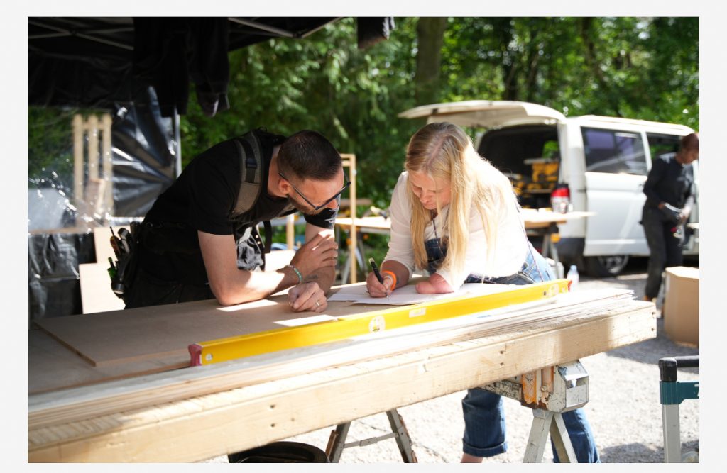 Emma is outside, leaning on a work table writing down plans for one of her pieces during the show. She stands next to one of the team carpenters.