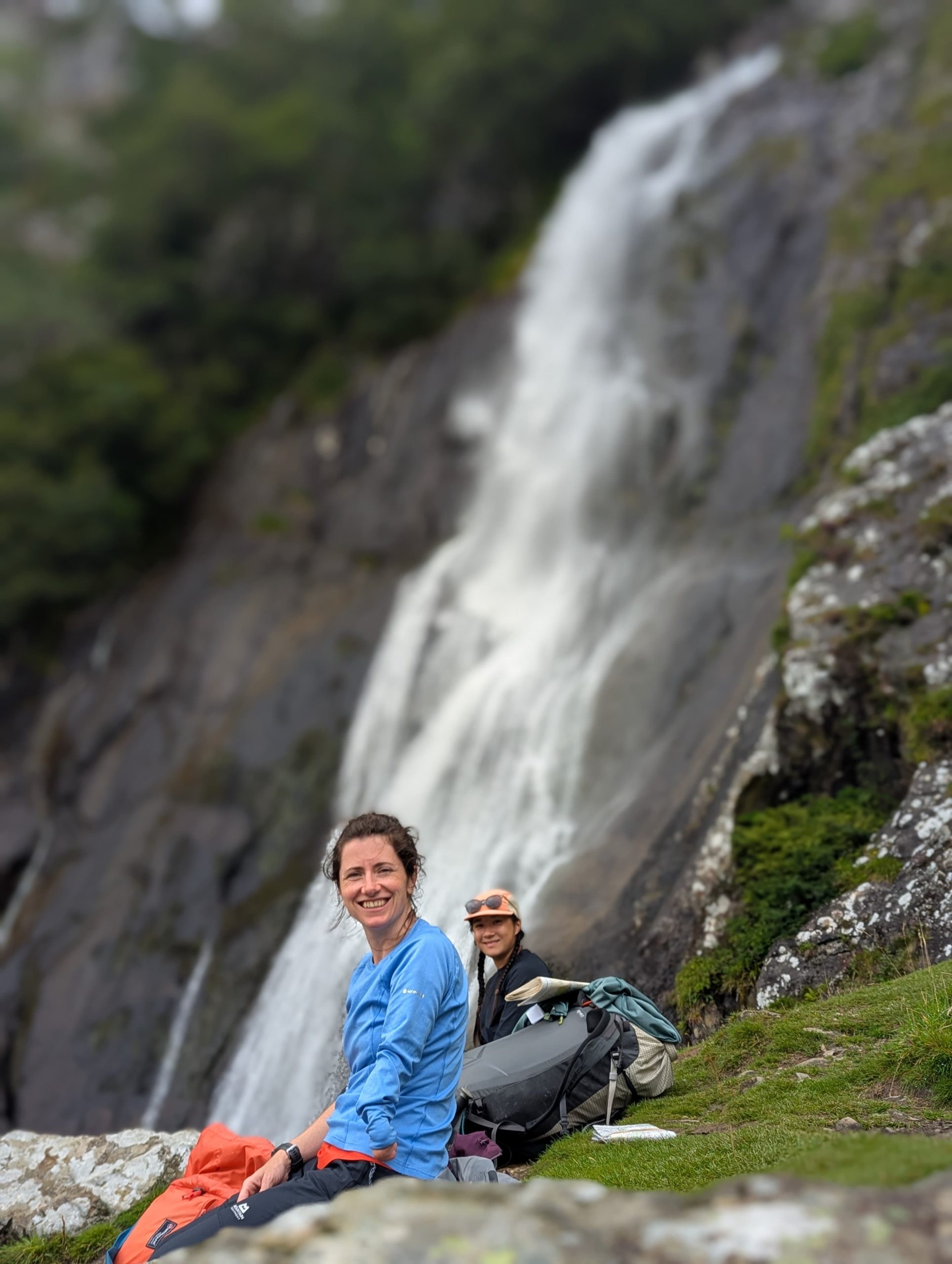 Max smiling and looking at the camera, sitting in front of a waterfall in North Wales during her mountain leader assessment week